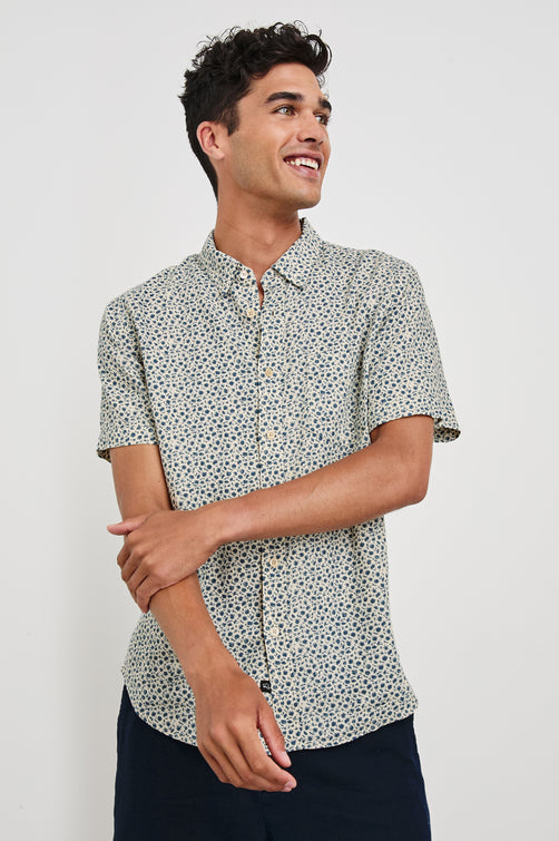 Carson linen short sleeve button down with a collar and blue botanical pattern in Autumn Daisy Ocean - front view on model, arms partially crossed looking away