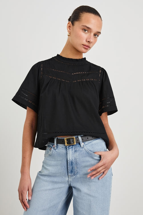 Front view of female model wearing our Daisy Top in black. This is a crewneck baby doll blouse, with ruffles at the collar. Styled with light wash denim and a simple belt.