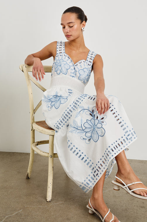 Model is sitting in a white chair wearing our sleeveless, Fawn Midi Dress with Blue Embroidery contrast and eyelet details. 