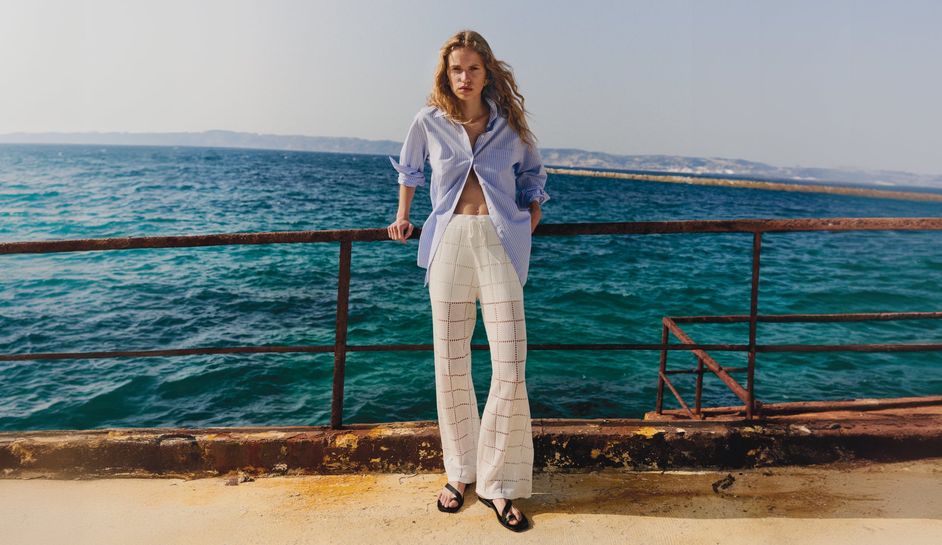 FRONT FULL BODY EDITORIAL IMAGE OF MODEL OUTSIDE BY THE OCEAN WEARING ELSA SHIRT IN OXFORD WHITE STRIPE AND MAJA PANTS IN WHITE