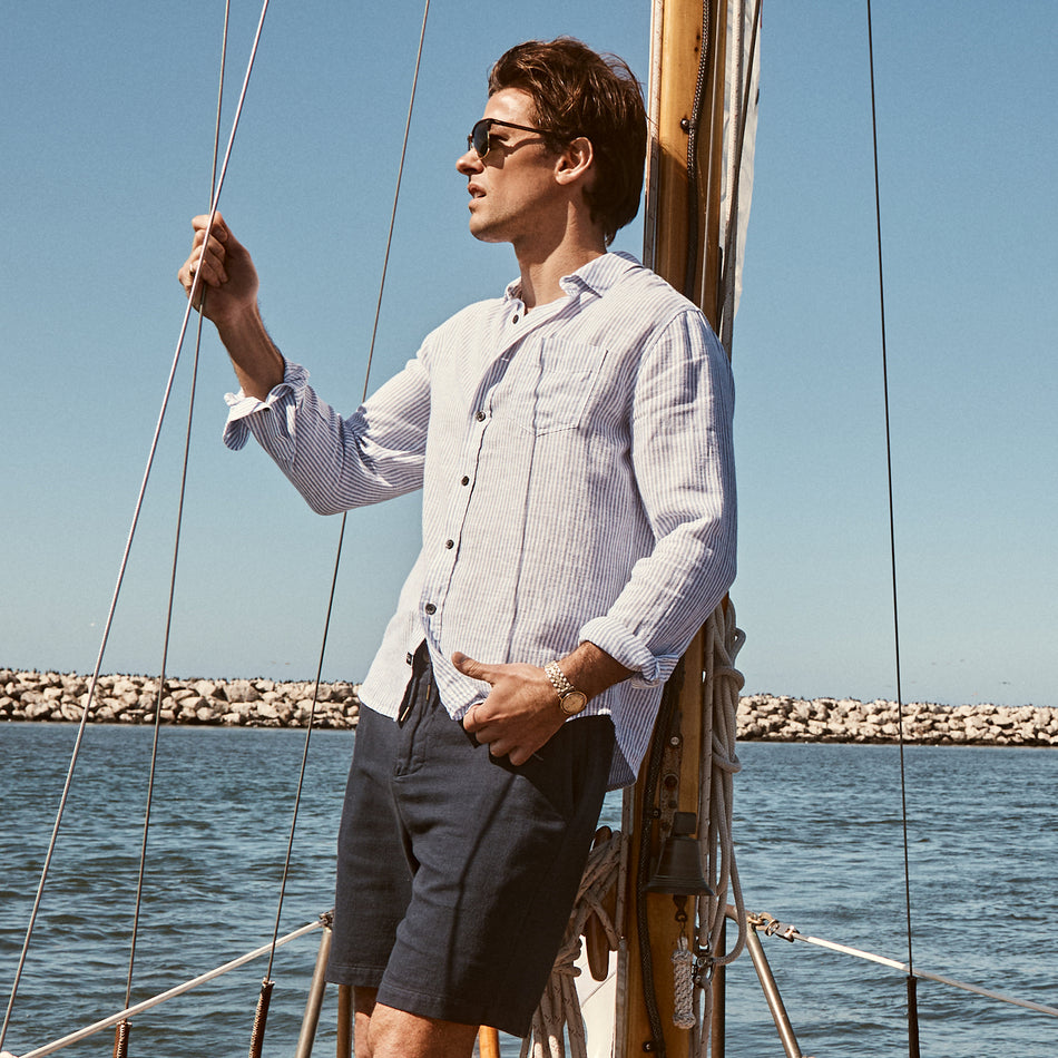 EDITORIAL IMAGE OF MODEL STANDING ON A SAILBOAT WEARING CONNOR SHIRT IN DENIM WHITE RAILROAD AND ARCHER SHORTS IN DEEP MERIDIAN.