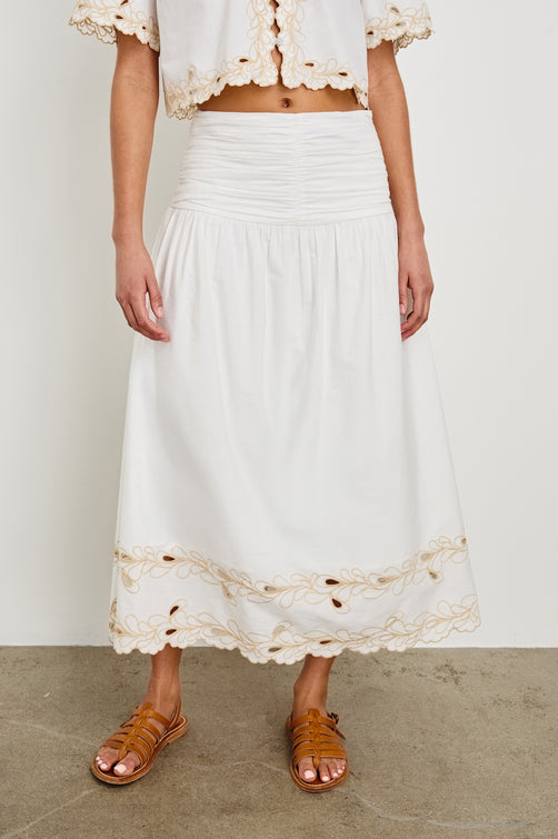 Detail image of model wearing a white, cotton poplin midi skirt with a lightly gathered waist that cinches and drops at the hips. Details include beige embroidered eyelets at the hem. Worn with brown sandals.